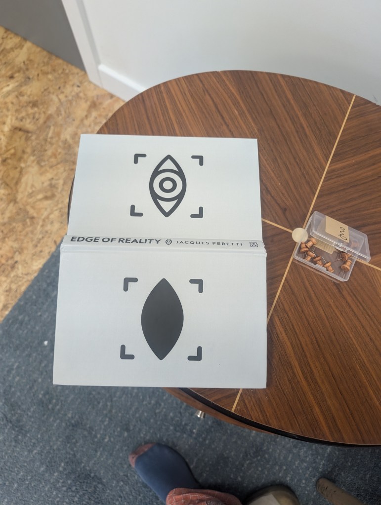 A book lies open on a table with the front and back cover turned upwards to towards us. The book is titled "Edge of Reality" by Jaques Peretti. There is a black and white eye shaped icon inside a square on the front cover, and the same icon on the back cover, but with the eye shape shaded in black.
